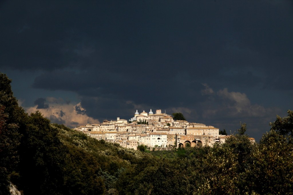 A picturesque hilltop village under a dark, cloudy sky, showcasing stone buildings and a prominent church dome.