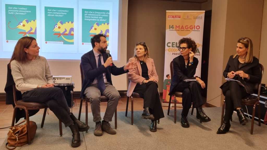 A panel discussion featuring four individuals sitting on chairs, with a man speaking into a microphone. Behind them are colorful posters related to health topics. The setting appears to be a conference or seminar.