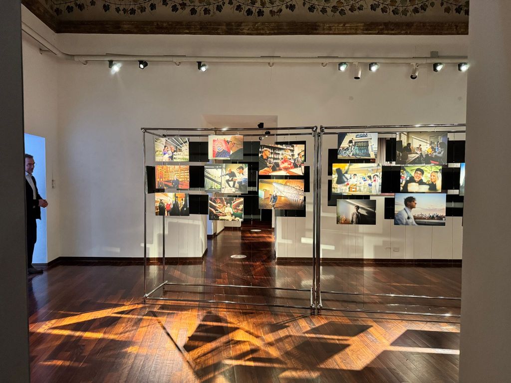 An art exhibition space featuring a display of photographs on a metal frame, with various images showcasing people and scenes. The inviting room has wooden floors and soft lighting, with a person in a suit visible in the background.
