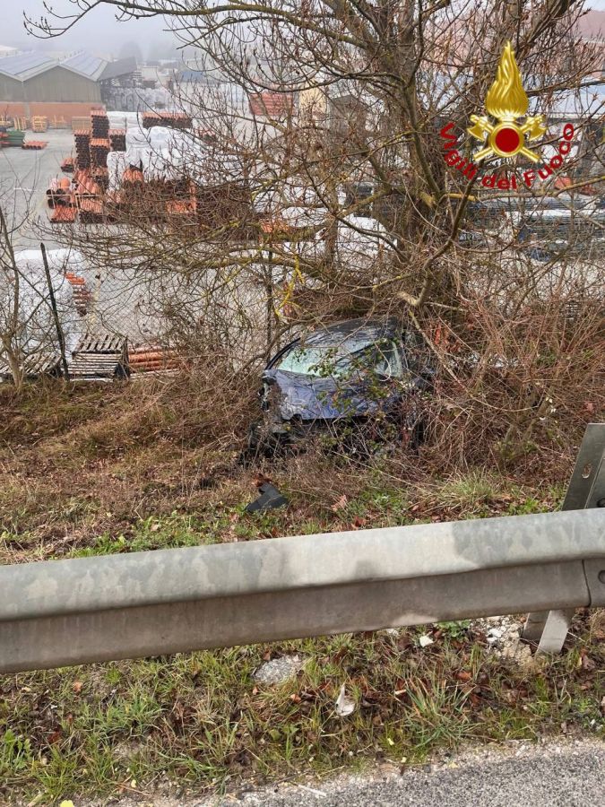 A car partially hidden in bushes near a roadside barrier, with a foggy background and stacks of materials in a storage area.