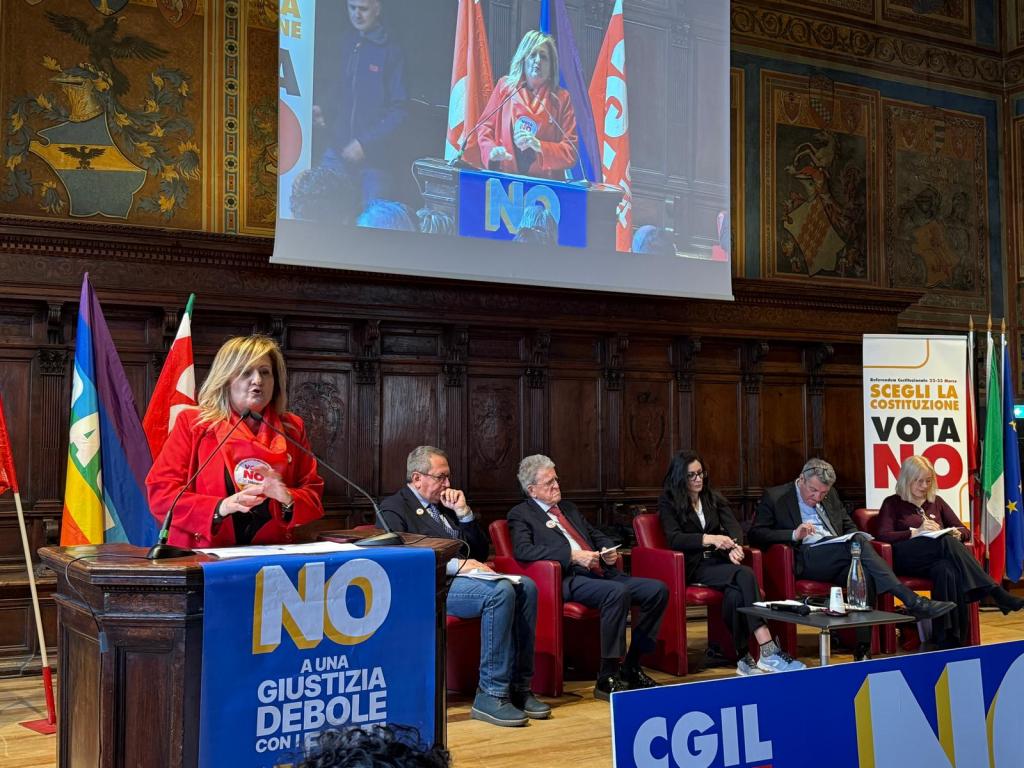 A public speaking event featuring a woman in a red jacket addressing an audience, with a large sign reading 'NO' on the podium. Behind her, there are flags and a screen displaying her image. Several seated speakers are visible in the foreground.