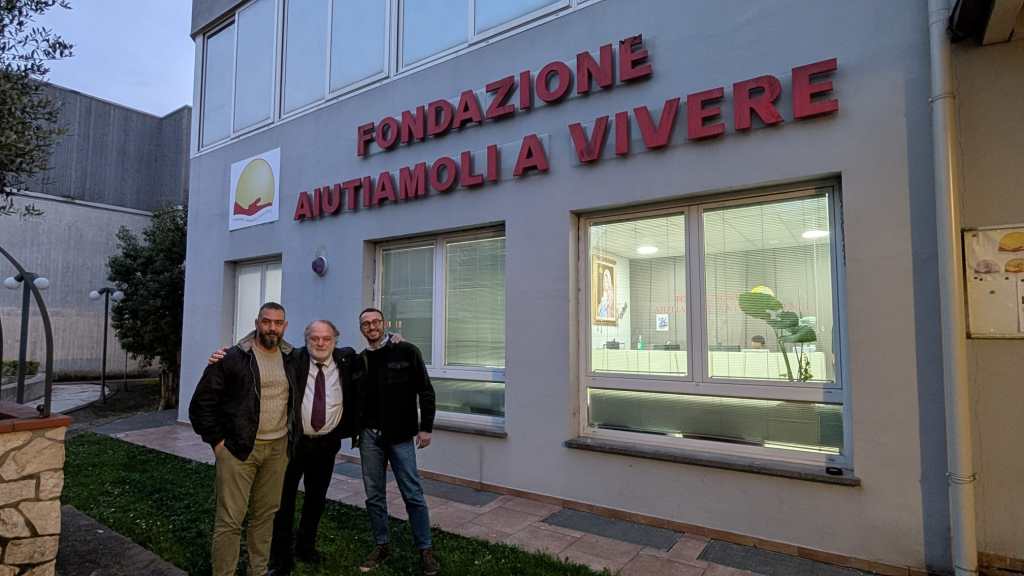 Three individuals standing in front of a building with the sign 'Fondazione Aiutiamoli a Vivere.' The building features large windows and greenery in the surroundings.