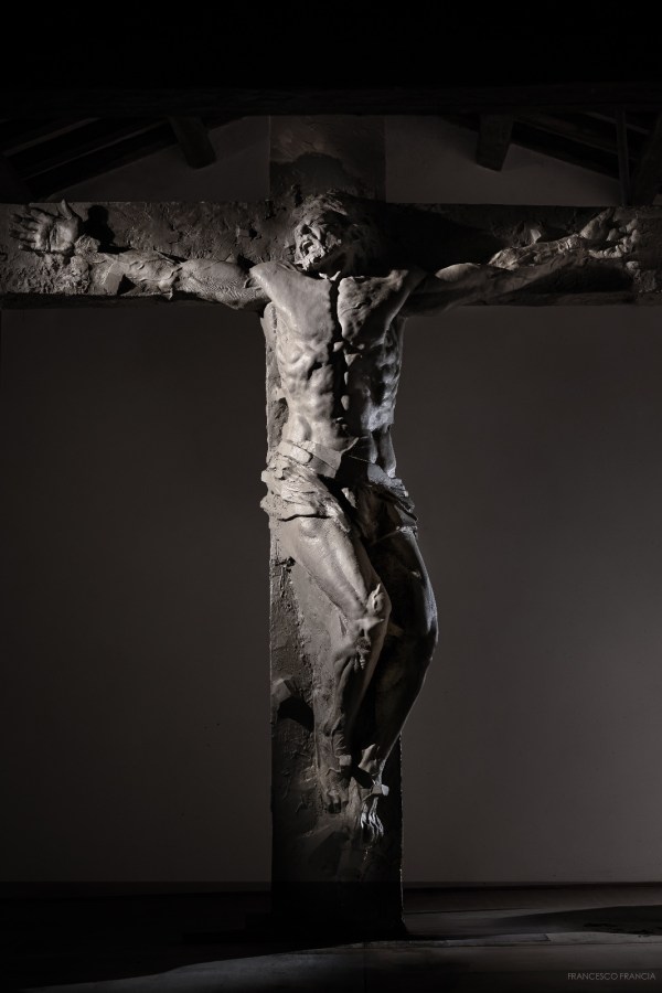 A dramatic sculpture of a crucified figure, emphasizing muscular details and emotional expression, set against a dark background.