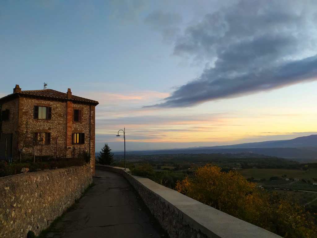A scenic view of a stone house alongside a winding path at sunset, with rolling hills and a colorful sky in the background.