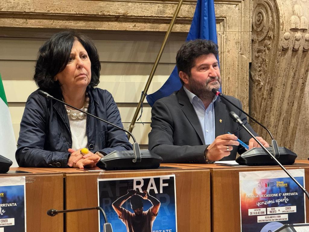 A man and a woman seated at a press conference table, with microphones in front of them. The woman is wearing a dark jacket and pearls, looking serious. The man is in a suit, holding a pen and speaking into a microphone, with a backdrop featuring flags and posters.