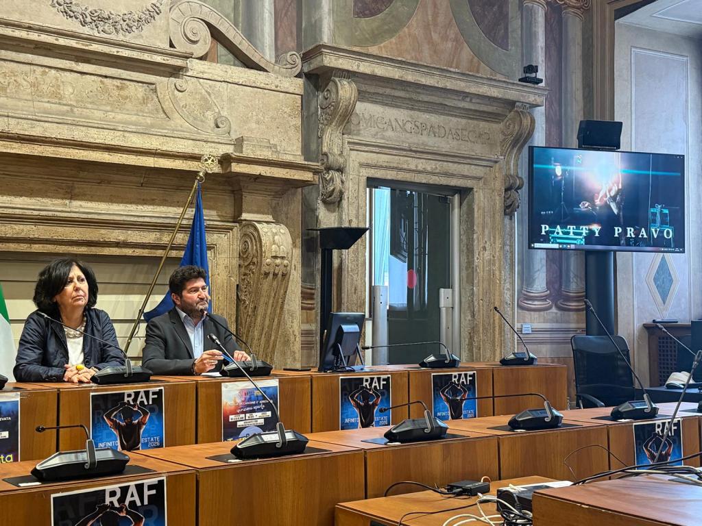 A press conference setting with two speakers at a wooden table, one woman and one man, in front of a backdrop featuring ornate decor. A screen displays the name 'Patty Pravo' along with promotional material for an event.