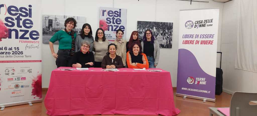 Group of women posing at a table during a feminist festival event, with banners and promotional materials in the background.