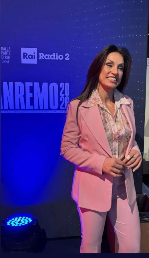 A woman in a pink suit stands in front of a blue background with text related to the Sanremo music festival, smiling for the camera.