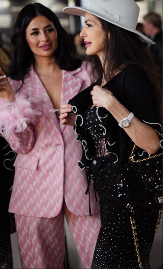 Two women engage in conversation at an event. One woman wears a pink patterned suit, while the other sports a black outfit adorned with white designs and a white hat.