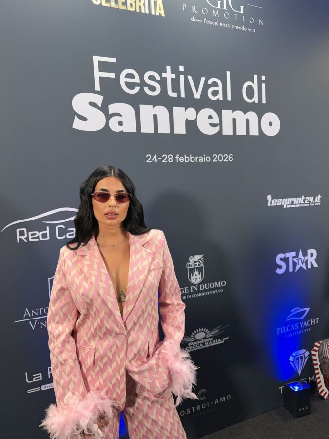 A person wearing a pink patterned suit with feather accents poses in front of a festival backdrop that reads 'Festival di Sanremo' and the dates '24-28 febbraio 2026'.