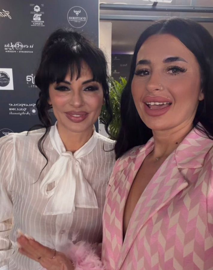 Two women posing for a selfie with cheerful expressions, one wearing a white blouse with a bow tie and the other in a pink patterned blazer.