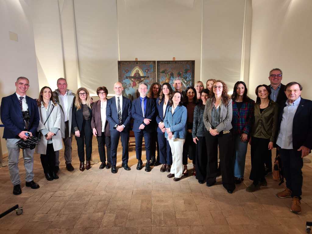 A group of diverse individuals posing together in a well-lit room, with a historic mural or artwork in the background.