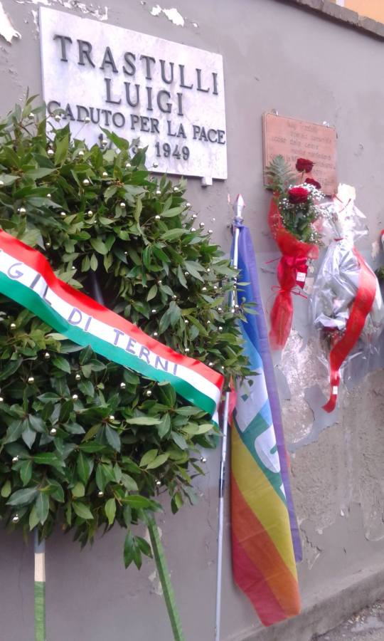 Memorial plaque for Luigi Trastulli, commemorating his sacrifice for peace in 1949, surrounded by a wreath and flags.