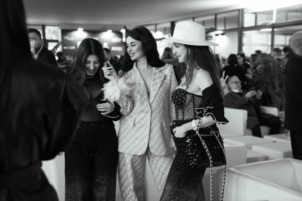 Three women in stylish outfits engaging in conversation at a social event. The setting is indoors, with soft lighting and other guests in the background.