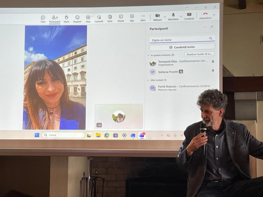 A virtual meeting setup with a woman on a screen, smiling in front of a building, while a man in a dark suit sits with a microphone, engaging with participants.