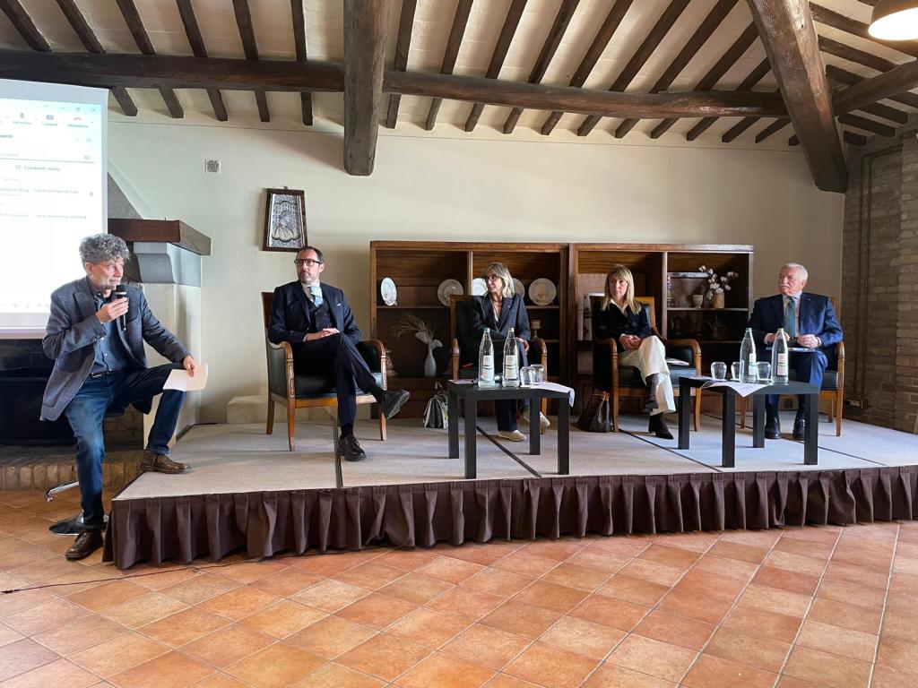 A panel discussion featuring four speakers seated at a low table, with one speaker standing and speaking into a microphone. The setting includes wooden beams and a rustic decor.