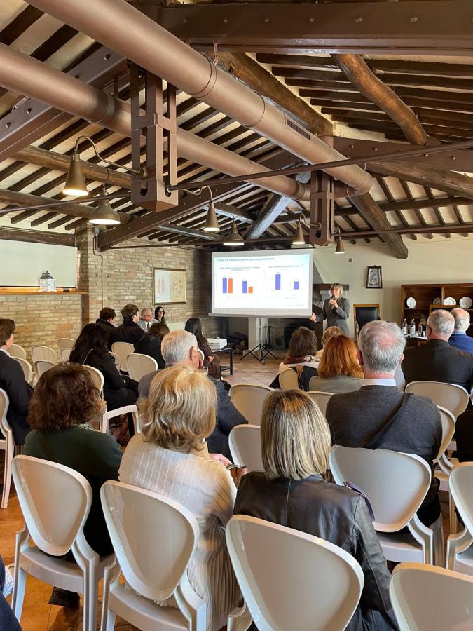 A speaker presenting data on a screen to an audience seated in a conference room with wooden beams and brick walls.
