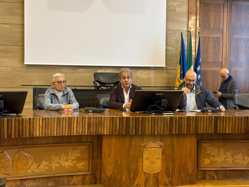 A meeting taking place in a conference room featuring three people seated at a wooden table, with one person speaking into a microphone while two others listen. Flags are displayed in the background.