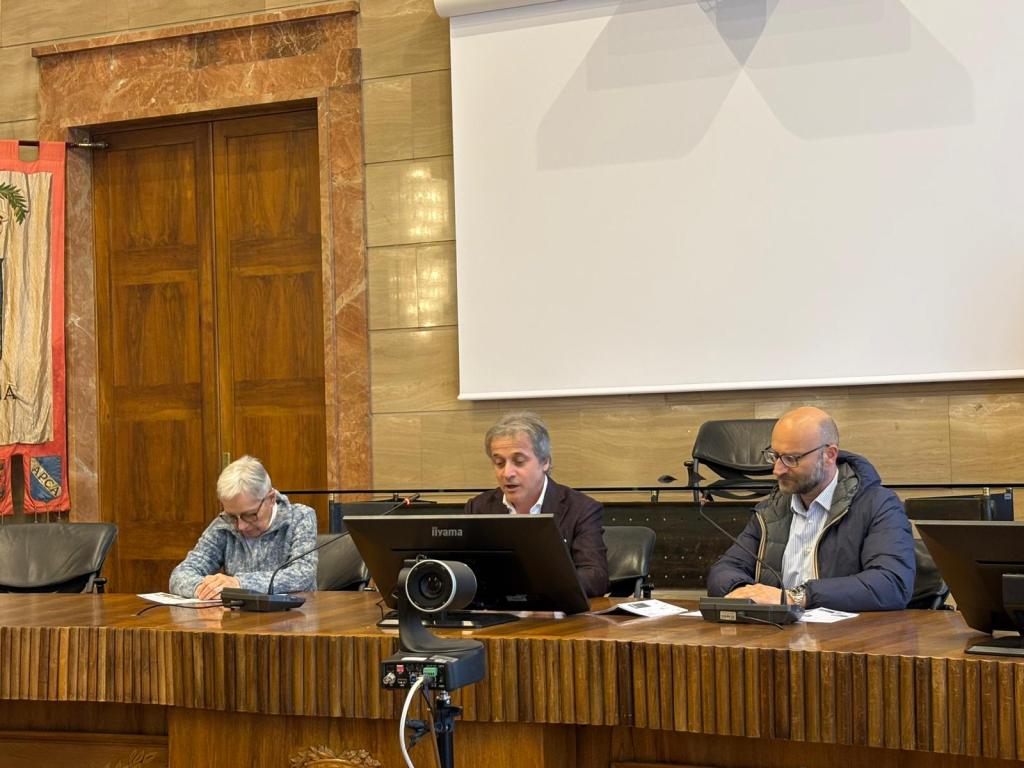 Three men seated at a long wooden table during a meeting, with a large screen in the background and wooden paneling on the walls.